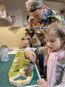 Córka z tatą w pracowni, wspólnie malują na zielono drewnianą planszę do gry w mankalę