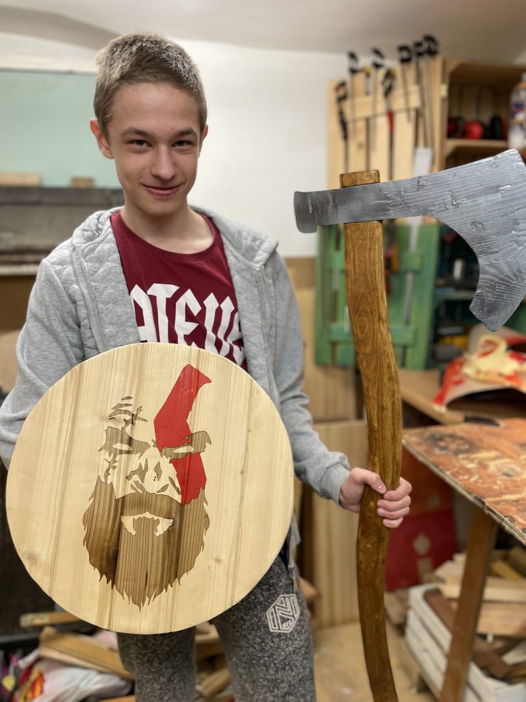 Nastolatek ubrany w tshirt, w warsztacie stolarskim, na tle narzędzi, trzymający drewniany topór